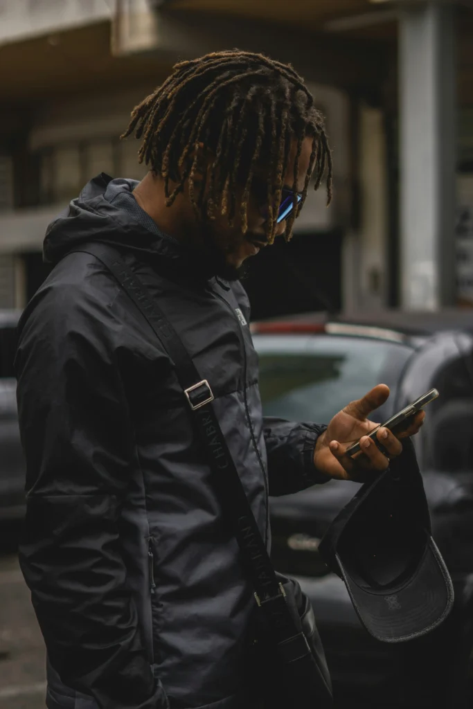 Person with dreadlocks wearing gray hoodie and backpack looking down at smartphone while standing on city street with cars in background.